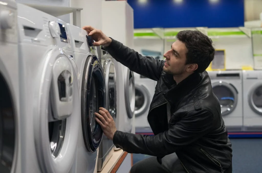 A man in a black jacket looking at a washing machine while contemplating appliance repair service options.